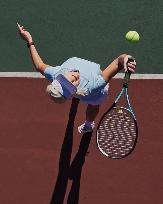 Overhead view: Woman in blue top, white visor & skirt on a green & brown tennis court. She holds a blue racket with a smiley face, looking up at a yellow tennis ball in mid-air. Long shadows stretch from her.