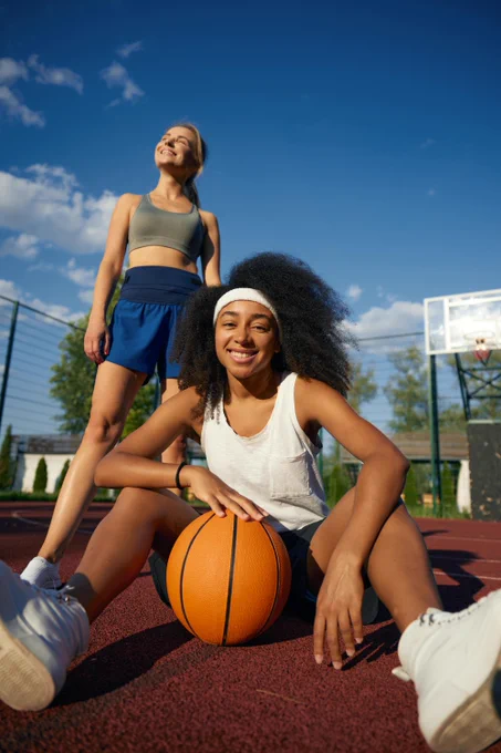 Two women on an outdoor basketball court. A Black woman with curly hair sits, smiling at the camera, hands on a basketball. Behind her, a white woman stands, eyes closed, smiling into the sun. Blue sky, basketball hoop.