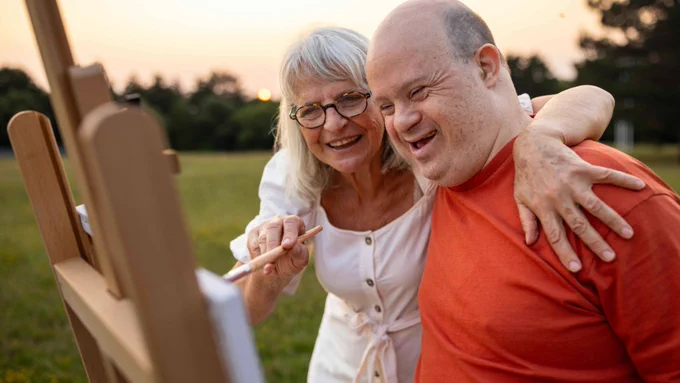 Two people are outdoors at an easel. One person is holding a paintbrush, and both appear to be enjoying the painting activity together. The setting is a grassy area with trees in the background.