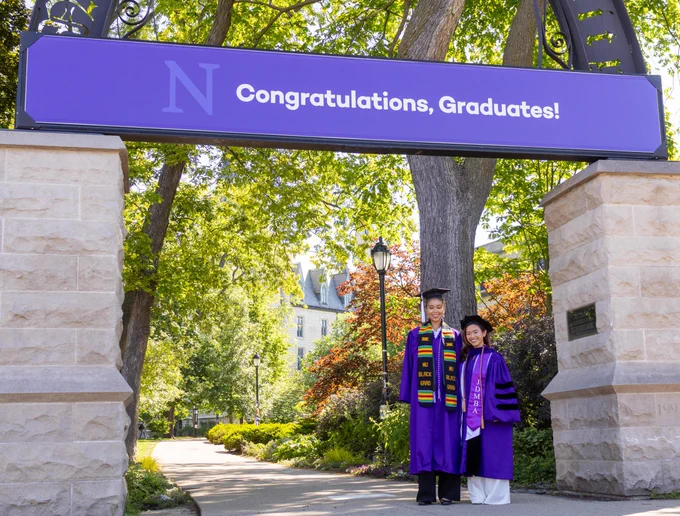 Sydney-Reese Harris (left) and Tram Nguyen (right) pose for a photo under Weber Arch while wearing their graduation attire. A sign above the arch reads \"Congratulations, Graduates!\"