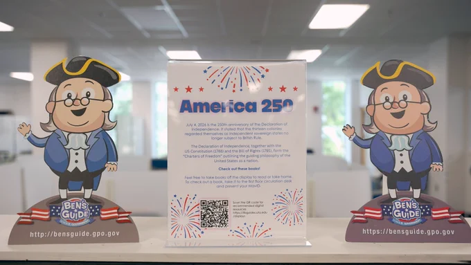 Pictured is a photo of to cardboard cutouts of Ben Franklin standing on each side of a sign that reads America 250th.