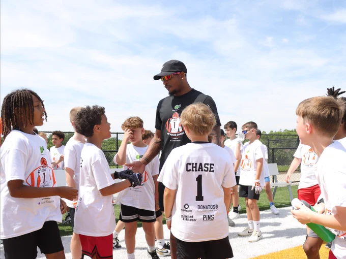 A person wearing sunglasses and a black T-shirt is interacting with a group of young athletes on a field. The young athletes are wearing white shirts with "CHASE" and the number "1" printed on them, indicating participation in a sports event. The sky is clear, suggesting a sunny day.
