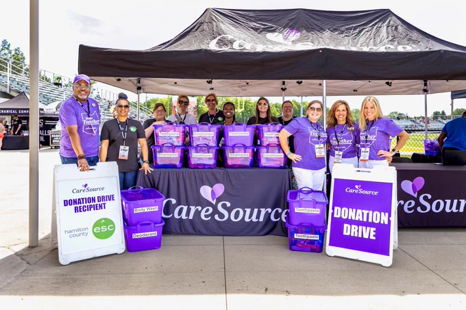 A group of people stands under a CareSource tent, participating in a donation drive event. They are holding and surrounding tables with purple CareSource boxes. Two signs read "CareSource Donation Drive." The event appears to be in an outdoor venue with a clear sky in the background.
