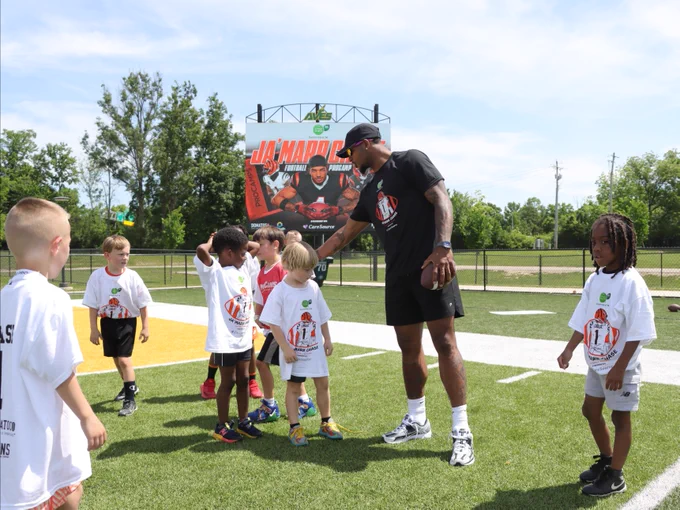 A group of children wearing sports shirts are on a football field, interacting with an adult holding a football. In the background. Trees and a fence are visible in the distance.
