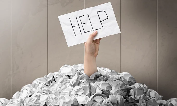 Hand sticking out from a pile of paper holding a note that says 'Help'