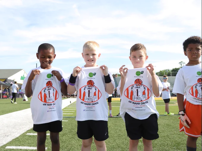 Four children on a sports field proudly hold t-shirts up. They smile towards the camera. Other participants and a sports venue are visible in the background.