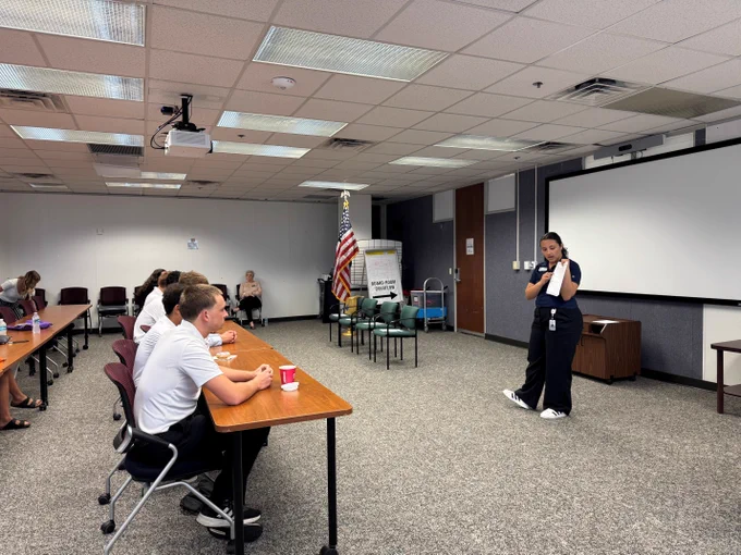 Female elections staff member giving a presentation and holding a ballot to a diverse group of attentive adults seated in a classroom.