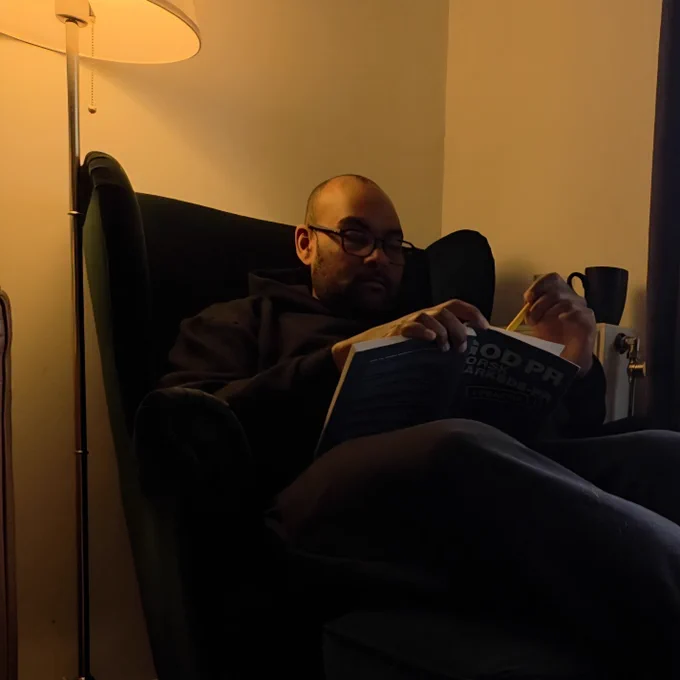A man sitting in a dark green chair under warm lamp light, reading and taking notes in a book with a pencil. The room feels calm, quiet and reflective.