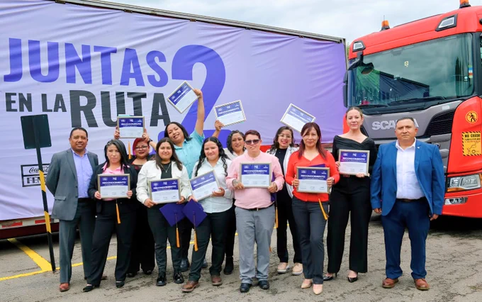Un grupo diverso de personas posa sonriente, sosteniendo certificados de reconocimiento frente a un gran camión rojo. Detrás, un cartel púrpura con el texto "Juntas en la Ruta". La escena parece ser parte de un evento de celebración.