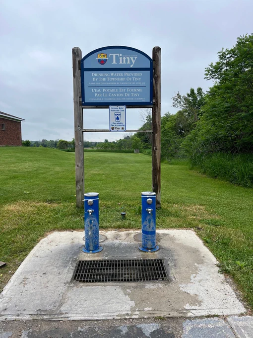 Two blue drinking water fountains stand on a concrete pad beneath a sign indicating water is provided by the Township of Tiny, also known as the Balm Beach Road water station.