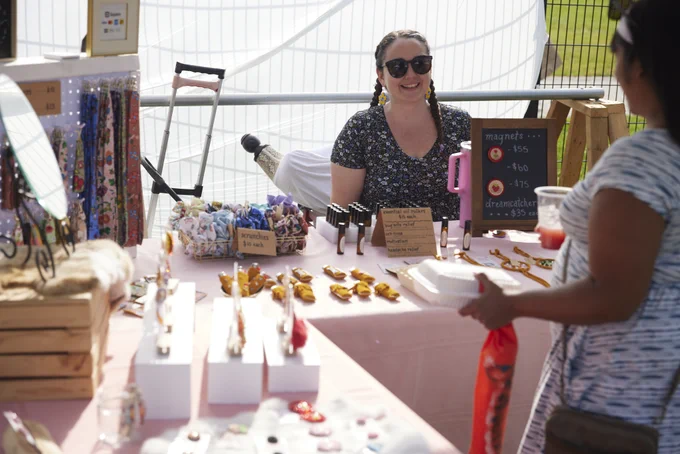 Vendor sitting behind a table displaying handmade jewelry and accessories at an outdoor market stall.