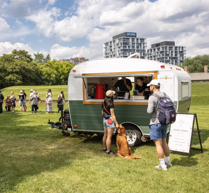 People ordering from a green and white food trailer in a sunny park with a dog sitting beside a woman at the window.