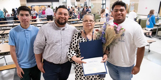 Group photo of four people standing in a school cafeteria, one holding a certificate and bouquet of flowers, celebrating an achievement.