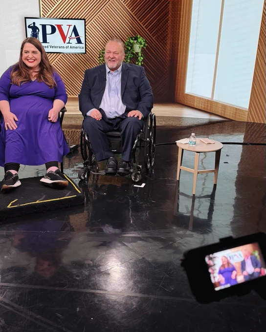 Maria Town, President and CEO of AAPD, and Tom Wheaton, National Treasurer of Paralyzed Veterans of America in a manual wheelchair, participate in a Satellite Media Tour discussing accessible air travel. The pair sit on a studio set in front of a screen displaying the PVA logo while a camera records the interview.