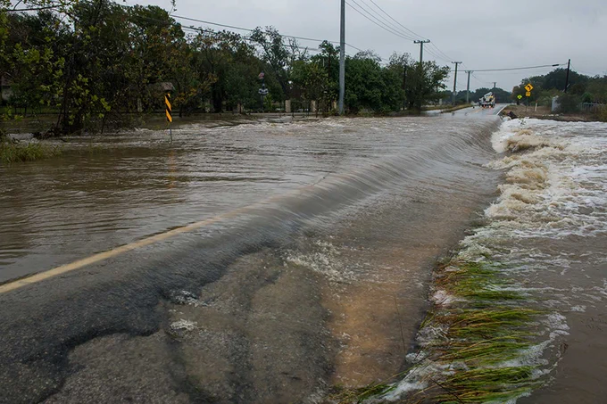 Floodwaters covering a rural road, making it impassable with strong currents visible on both sides.