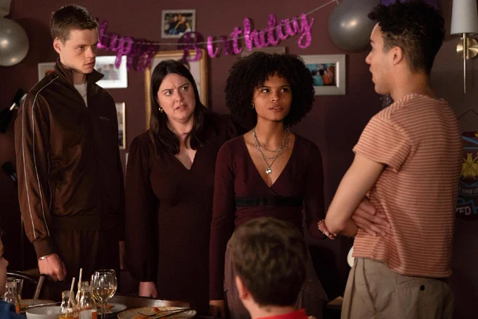 Oscar, Zoe, Jasmine and Josh stand in the Queen Vic living room room with a "Happy Birthday" banner on the wall in a scene from EastEnders. The table is set with glasses and plates.