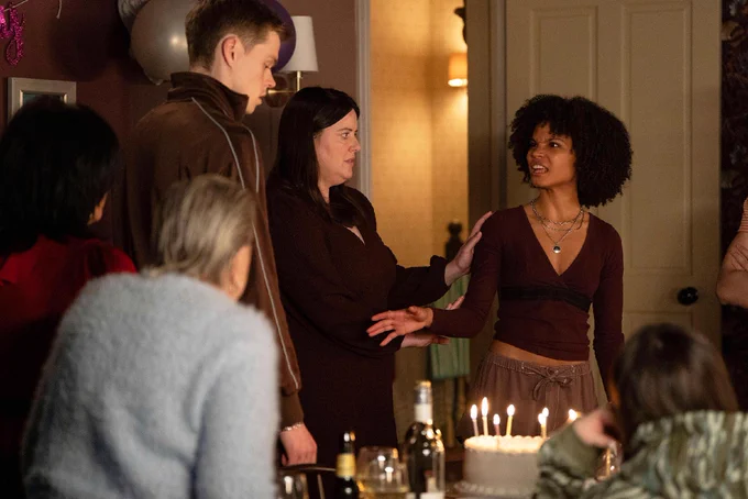 Kat, Mo, Oscar, Zoe and Jasmine standing around a table in the Queen Vic living room with a cake topped with lit candles in EastEnders. Bottles and glasses are visible on the table. Zoe is trying to hold Jasmine back as she angrily looks towards Oscar.