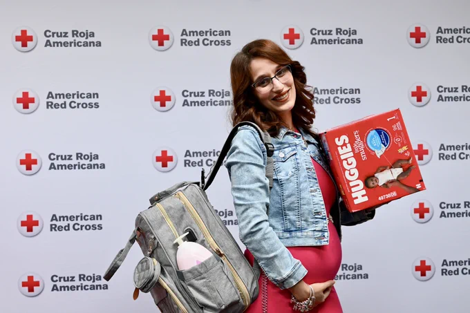 A person smiling stands in front of an American Red Cross and Cruz Roja Americana backdrop. They hold a box of Huggies diapers and carry a backpack with a baby bottle visible.