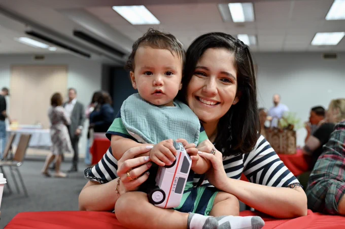 A person smiling and holding a child indoors. The child is holding a toy resembling a FedEx truck. The background includes people mingling and tables with red tablecloths.