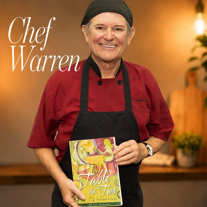 Chef Warren smiles warmly while holding his cookbook 'Table for Two.' The setting gives a professional yet inviting feel.