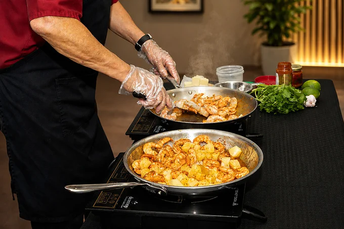 A chef wearing gloves prepares a seafood and potato dish in a professional kitchen setting. Ingredients like cilantro and limes are visible in the background.