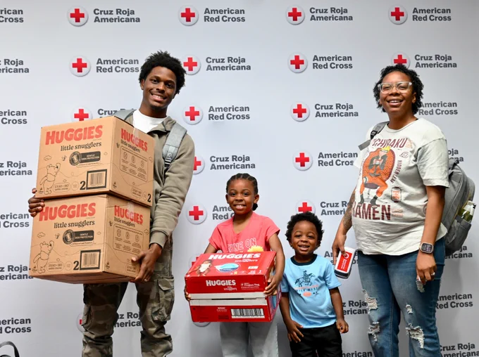A group of four individuals stand in front of a backdrop displaying the American Red Cross and Cruz Roja Americana logos. Two of the individuals are holding large boxes of Huggies diapers, while two children smile in the middle. The setting appears to be an event related to the organizations mentioned.