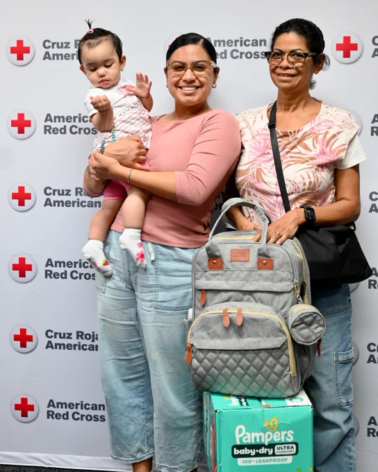 A group of three people are standing in front of a backdrop featuring the American Red Cross logo. One individual is holding a baby and smiling, while another is standing with a backpack and a box of Pampers diapers.