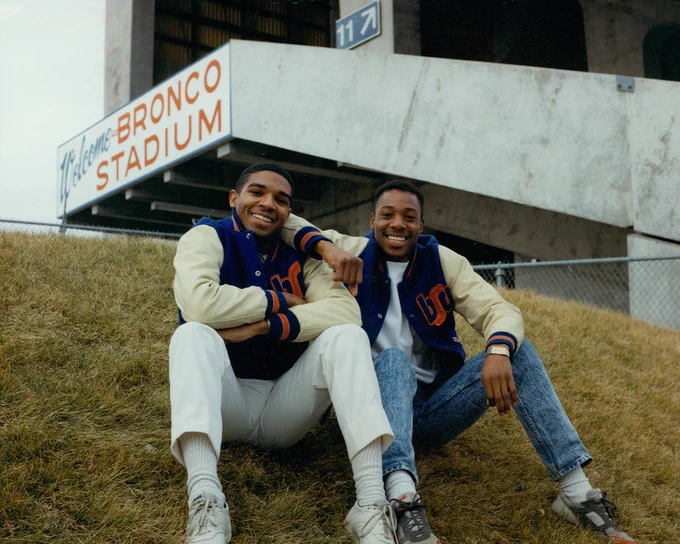 Two Boise State students from the 80s wearing letterman jackets smiling and sitting on a hillside near the “Bronco Stadium.”
