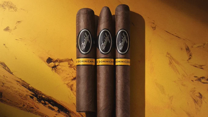 Three Davidoff cigars arranged vertically on a textured yellow-orange background. The cigars have dark wrappers and gold bands labeled "Puro Dominicano." - Cigars International