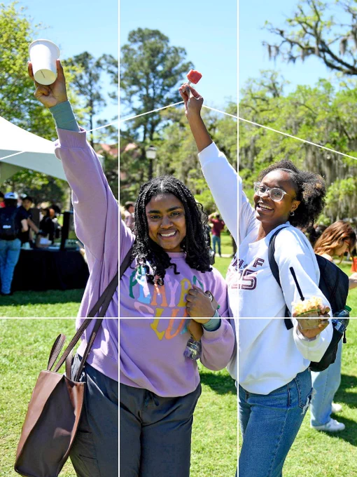Two people smiling and posing outdoors on Landis Green. One person is holding up a cup, the other has a popsicle. Trees and people are visible in the background. The classic "rule of thirds" lines are spaced on top of them, but adjusted in a quirky way to fit the subjects inside the center.