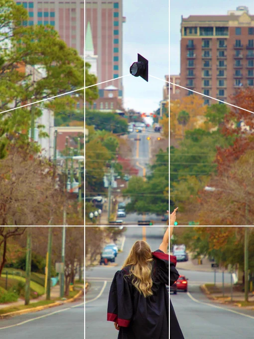 A person in a graduation gown is standing in the middle of a city street, tossing a graduation cap into the air. Tall buildings and a church steeple are visible in the background. The scene is framed by trees with autumn foliage. The classic "rule of thirds" lines are spaced on top of her, but adjusted in a quirky way to fit the graduation cap in the center frame.