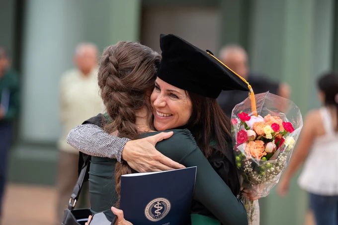Two individuals are embracing in a celebratory manner outdoors. One is wearing academic regalia, holding a bouquet of colorful flowers and a diploma with a medical symbol. The background is slightly blurred with a couple of people visible.