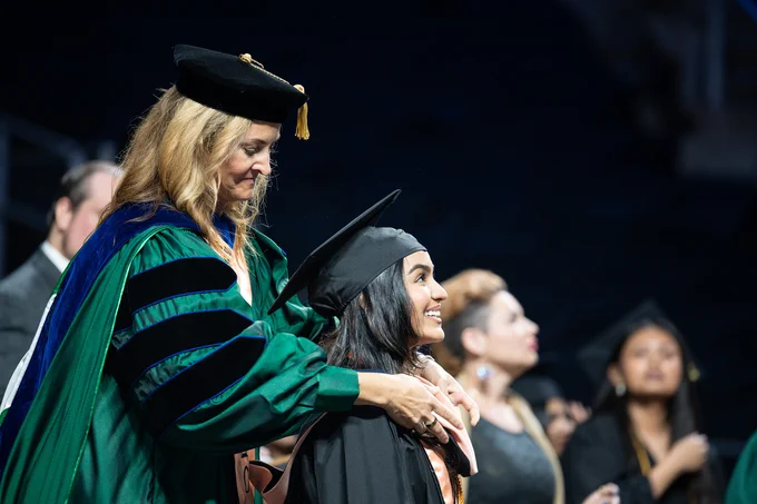 An individual in academic regalia places a stole over a graduate's shoulders during a ceremony, with others dressed in similar attire in the background.