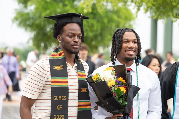Two people celebrate a graduation. One is wearing a graduation cap and a "Class of 2026" sash. The other holds a bouquet of colorful flowers. They're outdoors, surrounded by others and green trees.