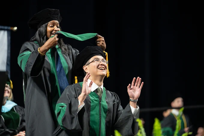A graduate in regalia is being hooded by another person on stage during a commencement ceremony. Other graduates are visible in the background, wearing similar robes and caps.