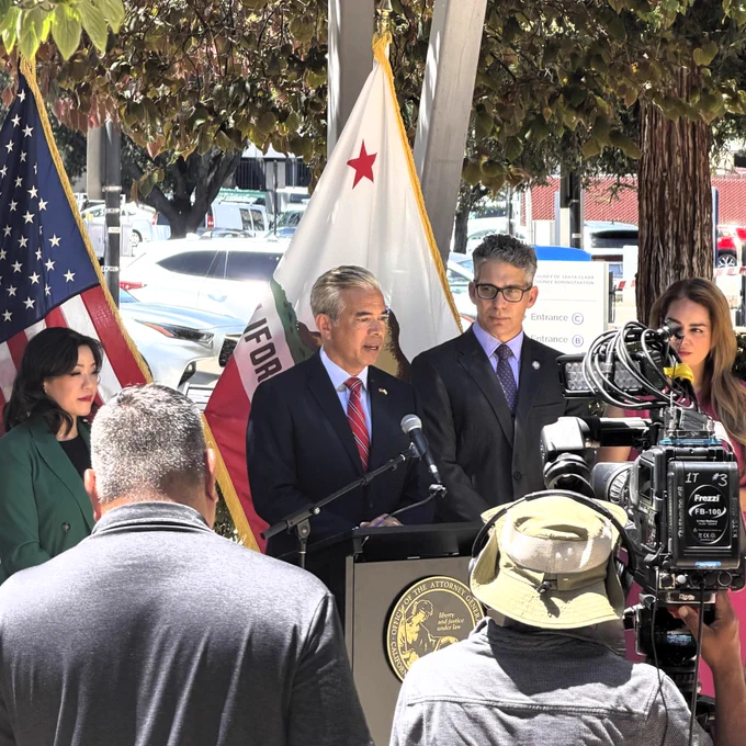 Attorney General Rob Bonta speaks  at a podium with a microphone. The U.S. Flag and California Flag are behind him as well as three people onlooking. There are news reporters, one with a camera in the foreground.
