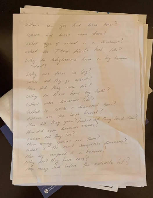 Photograph of a handwritten page containing a list of dinosaur questions submitted by kindergarten students ahead of a classroom presentation. Questions include topics such as where dinosaur bones come from, what T. rex fossils look like, why dinosaurs were so big, how they died, what is inside a dinosaur bone, why Ankylosaurus had a club tail, how many dinosaur species existed, and which dinosaur was the most dangerous. The page sits atop a stack of papers, highlighting the curiosity and imagination of young children preparing to learn about dinosaurs. 