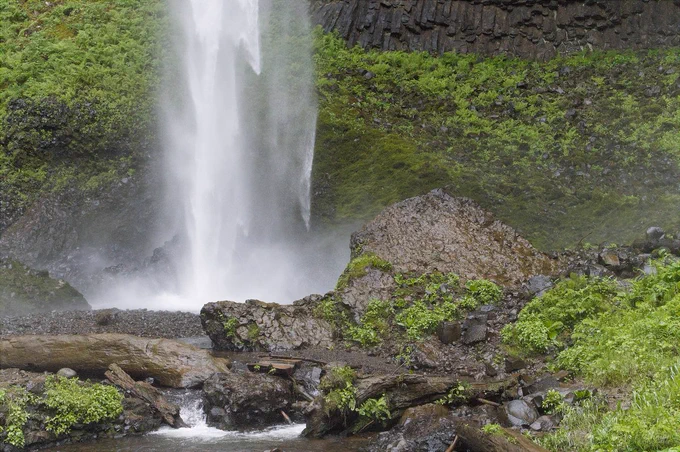 A close view near the base of a waterfall, with white water plunging down the left side of the frame into a misty pool below. Dark basalt rocks, wet logs, scattered stones, and bright green ferns fill the foreground and surrounding canyon walls. Mist softens the scene and adds a sense of movement around the falling water. The photo feels powerful, damp, and alive with the energy of the canyon.