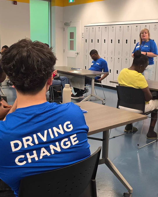 A seated volunteer is seen listening carefully to the other volunteer speaking during the session. The text on their t-shirt reads "Driving Change".