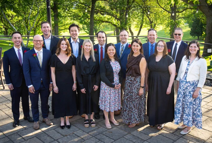 A group of fourteen physicians stand together outside on a paved area with trees and grass in the background, enjoying a sunny day. They are dressed in formal and semi-formal attire, smiling at the camera.