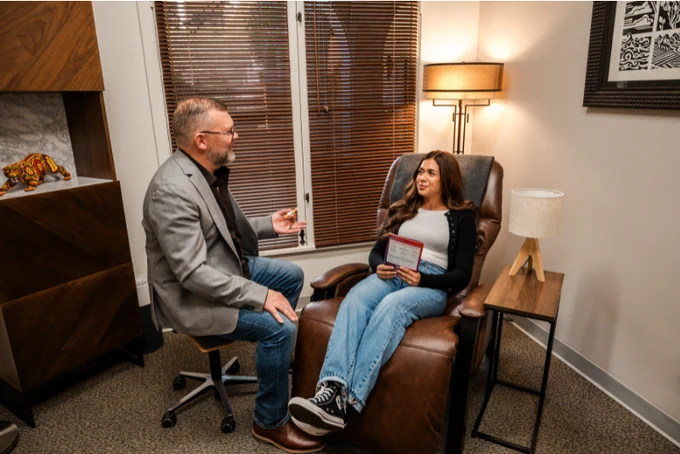 cozy-lit interventional psychiatry practice with woman sitting comofrtably in a leather chair while a male psychiatrist gestures and speaks to her