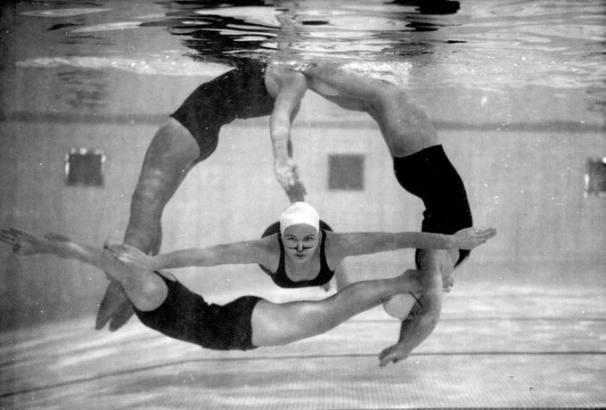 Black and white photograph of four synchronized swimmers underwater forming a circular pattern, bodies arched and arms extended, with pool tiles visible below