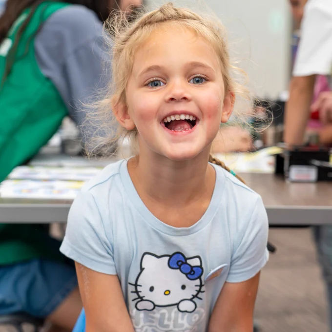 A child with a joyful expression in a library setting. In the background, others are engaged in activities around tables.
