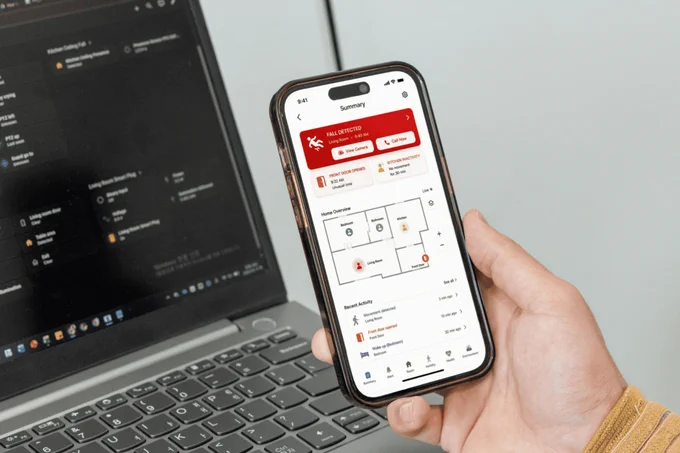 Person holding a smartphone displaying a security monitoring and fall detection app next to an open laptop on a desk.