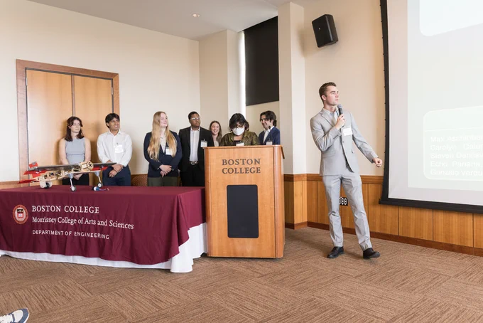 A group of individuals stand at a podium with a "Boston College" sign. The podium has a microphone, and one person speaks to an audience. A table with items, including a model plane, is to the side. The backdrop features a screen displaying text that is partially visible.