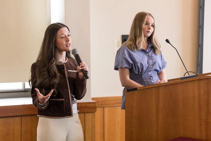 Two individuals are standing at a podium in a room. One person is speaking into a microphone, while the other stands behind the podium. The room has wooden paneling and a window with a shade drawn.