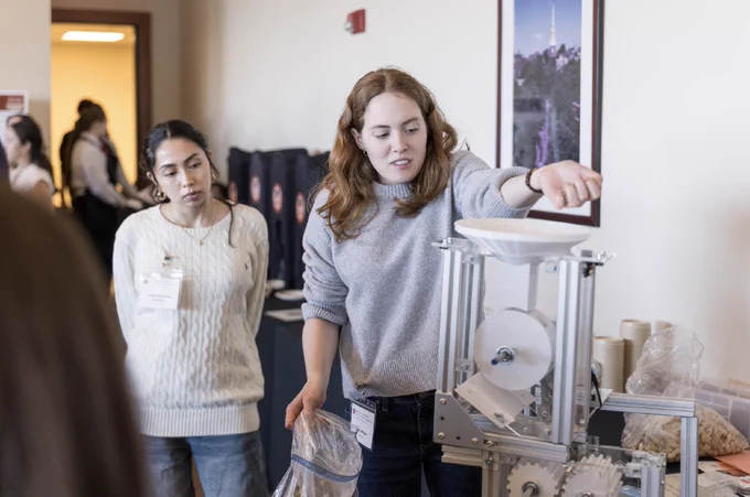 A person is demonstrating a mechanical device to an attentive individual at what appears to be an educational or scientific event. The room has visible posters and equipment, and additional people are present in the background.