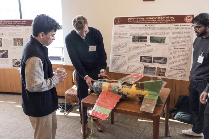 Three individuals are gathered around a table with a handmade model rocket. Two people are examining the rocket, while another holds a cup. Behind them are poster presentations with text and images, one titled "Democratizing Humanitarian Demining.