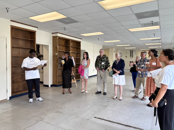 A group of nine people stand inside an empty commercial space with large front windows on the right side. The room has a drop‑ceiling with fluorescent lighting, unfinished flooring, and built‑in wooden shelving along the left wall. One person on the far left, wearing a white T‑shirt, holds a clipboard or paper and addresses the group. The others stand in a loose line facing toward the speaker, holding papers or small items. An easel with a poster is positioned near the center-left of the room. 