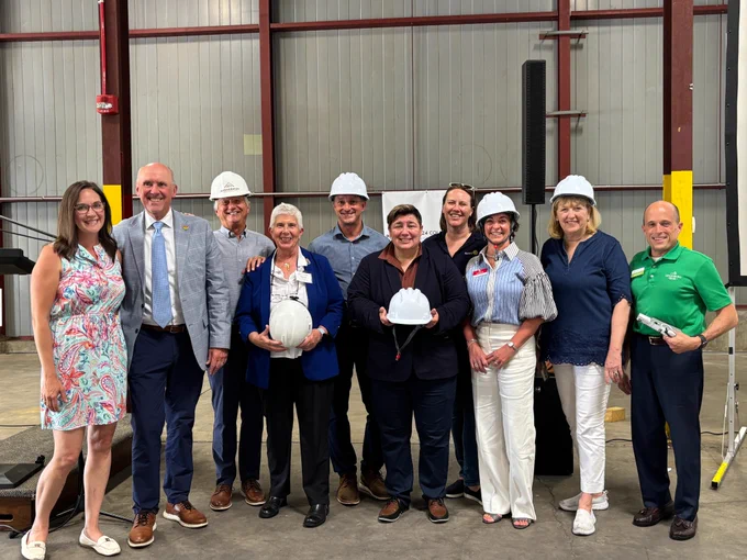 A group of twelve people stands closely together inside an industrial warehouse space with tall metal walls, exposed steel beams, and a concrete floor. Several members of the group wear white construction hard hats, and one individual at the center holds an additional hard hat in both hands. The group is dressed in a mix of business and casual clothing, including jackets, button‑down shirts, patterned dresses, and slacks. Behind the group is a banner attached to the wall, partially visible, along with yellow safety padding on the steel beams.